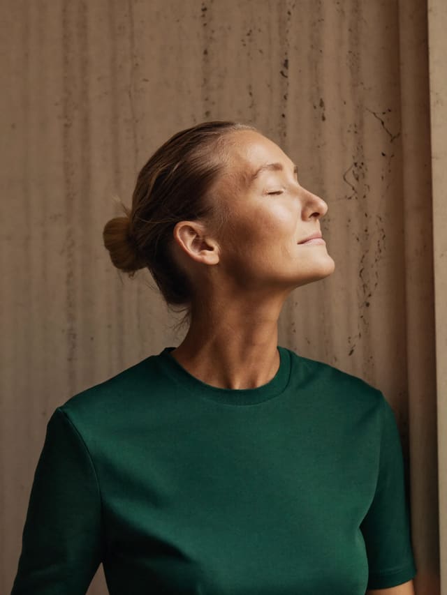 Model in a green T-shirt in warm light, eyes closed, against a travertine wall