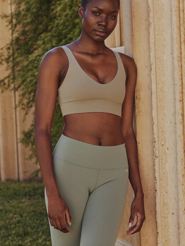 Model in a taupe sports top and sage leggings beside a stone column